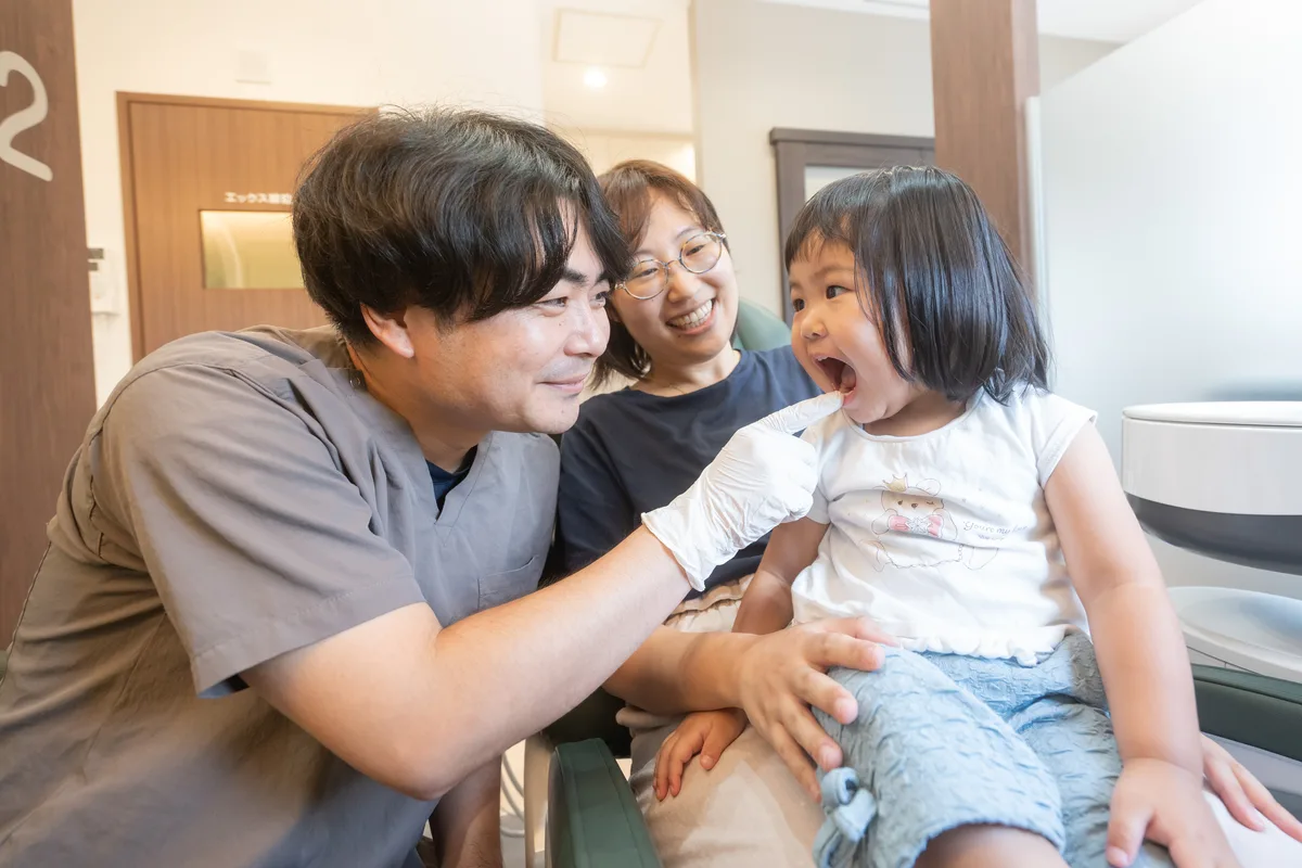 保護者のひざに座った女の子の口の中を歯科医師がのぞき込む様子